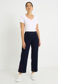 Pantalones anchos de mezclilla oscura con cintura alta y detalles de botones, combinados con una camiseta blanca de algodón de manga corta y zapatillas blancas.