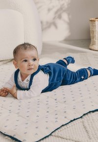 Baba kék bársonyos overálban és fehér pólóban, egy mintás takarón fekszik. A háttérben lágy textúrák és természetes fény látható.