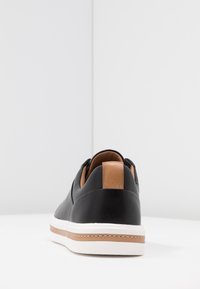 Sneaker en cuir noir avec une accent en tan sur le talon, semelle en caoutchouc blanche et couture contrastante. Texture lisse et design élégant.