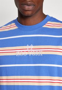 Camiseta de algodón azul con rayas horizontales en beige, rojo y blanco. Presenta un logotipo bordado de "ZOO YORK" en blanco en el pecho.