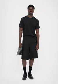 Junger Mann steht, trägt ein schwarzes T-Shirt, knielange Shorts, schwarze Socken und Schuhe und hält eine schwarze Clutch-Tasche in der rechten Hand.