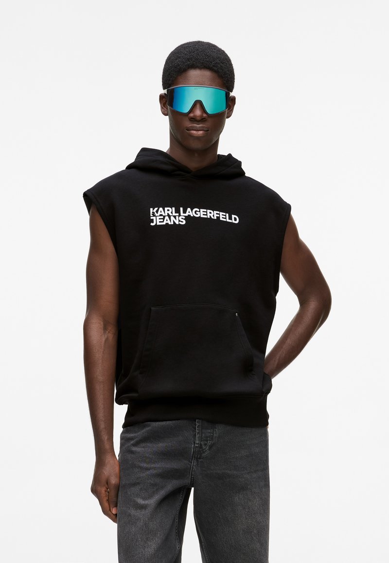 Felpa nera senza maniche con tasca frontale e testo bianco "KARL LAGERFELD JEANS". Modello che indossa occhiali da sole riflettenti blu con design piatto.