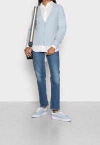 Ljusblå stickad cardigan med V-ringning, knäppning fram, lagen över en vit skjorta, i kombination med blå jeans och ljusblå sneakers.