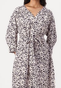 Puvun napitettava mekko, joka on valmistettu kevyestä kankaasta; siinä on musta ja kerma beige abstrakti kuvio, V-pääntie, pitkät hihat ja sidottu vyötärö.