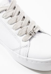 White sneaker with thick beige laces and a small silver "TH" logo detail near the bottom lace hole.