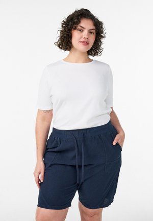 Vrouw met krullend haar, gekleed in een wit shirt met korte mouwen en marineblauwe shorts met trekkoord, staand met één hand in de zak.