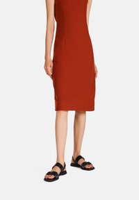 Sandali neri con un design floreale decorato e cinturini regolabili, indossati con un vestito aderente color ruggine a lunghezza ginocchio. Texture liscia.