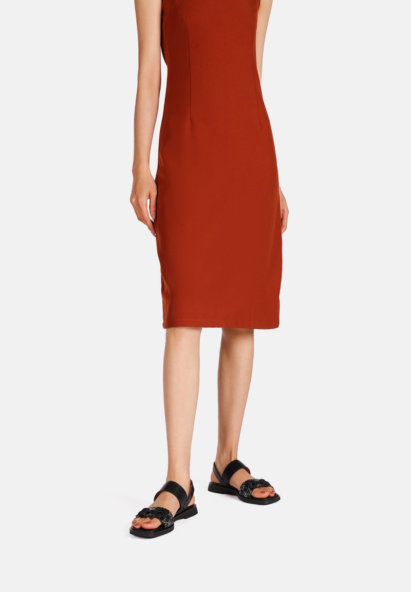 Sandali neri con un design floreale decorato e cinturini regolabili, indossati con un vestito aderente color ruggine a lunghezza ginocchio. Texture liscia.