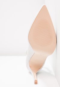 Chaussure à talon haut en daim beige avec un bout pointu et un talon aiguille élancé. La semelle est lisse avec un motif texturé subtil.