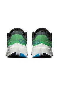 Des chaussures de course avec une tige noire accentuée de vert, une semelle blanche et des détails bleus. Le texte sur le côté indique "INGÉNIERIE SUISSE" et "ON."