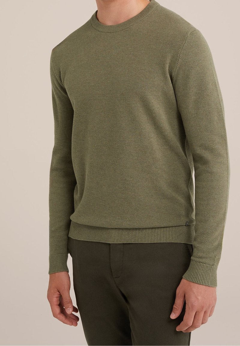 Homme portant un pull texturé vert olive à manches longues et un pantalon vert foncé, debout devant un fond clair uni.