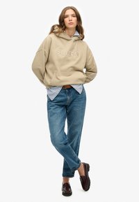 Beige hoodie met het logo "Superdry", gelaagd over een blauw-witte gestreepte shirt, gecombineerd met blauwe denim jeans en donkere instappers.