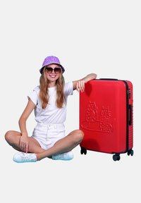 Red hard-shell suitcase with embossed cat design, black zipper, and four wheels. Model wears white shirt, shorts, and purple bucket hat.