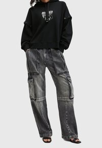 Černá mikina s kapucí a grafickým designem na hrudi, v kombinaci se šedými cargo kalhotami s několika kapsami a kontrastním šitím.