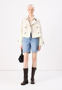Giacca cropped beige chiaro con bottoni doppi, pantaloni shorts in denim blu, stivali marroni alla coscia e una borsa nera con dettagli floreali.