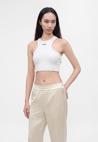 Jeune femme aux longs cheveux noirs portant un top court blanc avec le texte "OFF" et un pantalon beige à taille élastique, debout devant un fond uni.