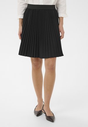 Naine, kes kannab musta voltidega seelikut, valget pluusi rüüskeerustega varrukatega ning tumedaid terava ninaga slingback-kingi, seisab valgel taustal.