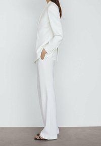 Blazer blanc ajusté avec une fermeture à un bouton, associé à un pantalon ample assorti. Texture lisse et design minimaliste.