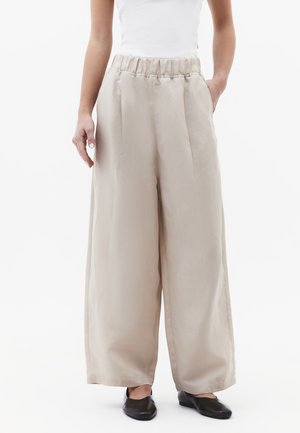 Kvinde iført beige bukser med vidde ben og elastisk talje samt sorte flade sko, hænder afslappet langs siderne mod hvid baggrund.