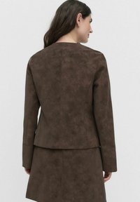 Femme avec de longs cheveux noirs portant une veste ajustée en daim marron et une jupe, vue de dos sur un fond uni.