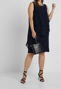 Mörkblå lagerklänning med en pärlad halsringning, kombinerad med en svart handväska och svarta sandaler med remmar. Slät tyg med en flödande silhuett.