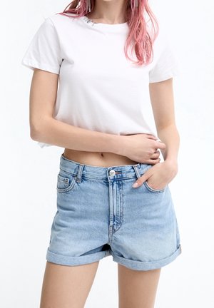 Młoda kobieta z różowymi włosami, ubrana w biały krótki T-shirt i jasnoniebieskie szorty jeansowe z wysokim stanem i podwiniętymi mankietami.