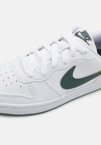Bela trenirka z gladkim usnjenim zgornjim delom, ki ima zeleno Nike potezo, perforirano prednjo škatlo in bele vezalke. Teksturiran podplat.