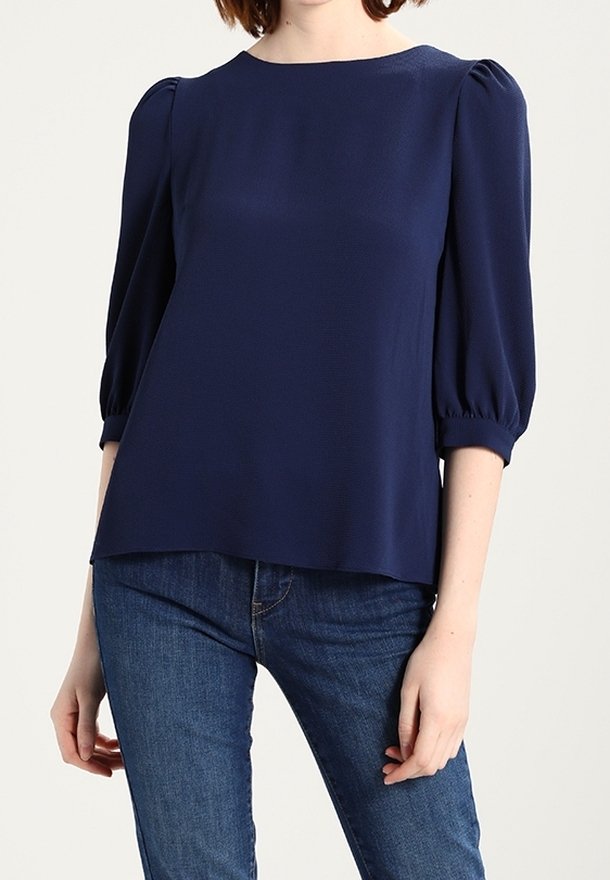 Femme portant un chemisier bleu marine à manches trois-quarts bouffantes, associé à un jean bleu sur un fond uni.