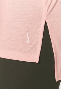 T-shirt de sport rose clair à manches courtes, avec une fente latérale et un logo Nike blanc positionné près de l'ourlet, associé à des leggings vert foncé.