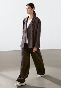 Brown double-breasted jacket with large buttons, worn over a white top and paired with matching wide-leg trousers. White shoes complete the outfit.