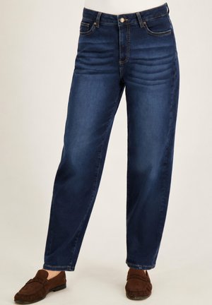 Blauwe rechte jeans met voorzakken en knoopsluiting, gedragen met bruine suède loafers op een lichte achtergrond.