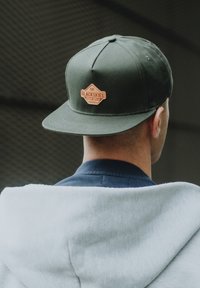 Cappello verde con visiera piatta, caratterizzato da una toppa ricamata marrone con la scritta "BLACKSKIES" sul retro. Felpa con cappuccio grigia visibile sotto.