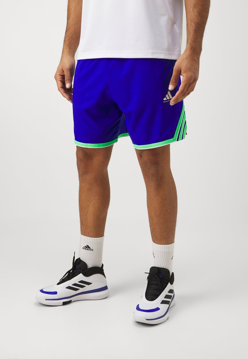 Königsblaue Sportshorts mit grünen Akzenten, die drei schwarze Streifen an den Seiten aufweisen. Kombiniert mit schwarz-weißen Turnschuhen.