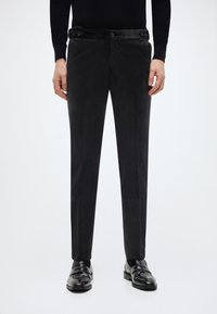 Schwarze, maßgeschneiderte Hose mit glattem, mattem Finish, ausgestattet mit einem taillierten Bund mit Knopf und geradem Bein. Kombiniert mit schwarzen Slippern.