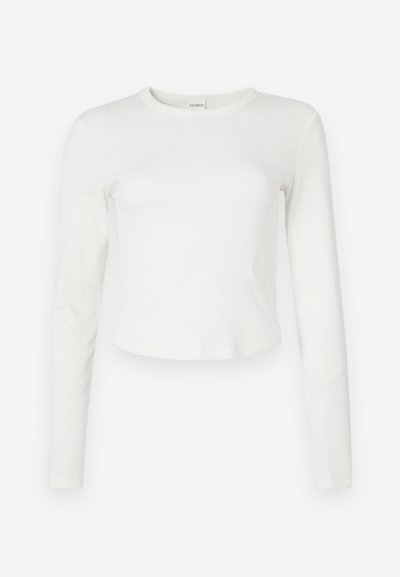 White long-sleeve ribbed top with a round neckline and cropped hem. Soft texture and minimalistic design, featuring a small logo on the side.