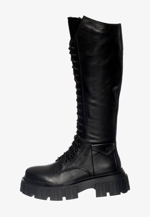 Bottes noires montantes à lacets avec semelles crantées épaisses et fermetures éclair latérales, portées sur des jambes nues.