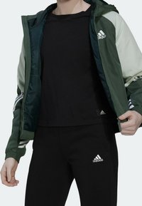 Vihreä ja vaaleanharmaa vetoketjullinen huppari, jossa valkoiset Adidas-raita, yhdistetty mustaan T-paitaan ja mustiin housuihin, molemmissa logolla.