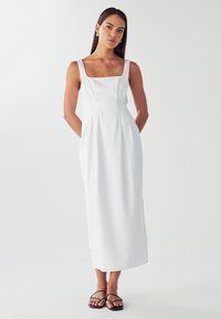 Biely šaty s štvorcovým výstrihom, priliehavým korzetom, splývavou sukňou a bočnými vreckami; doplnené tenkými čiernymi remienkovými sandálmi.