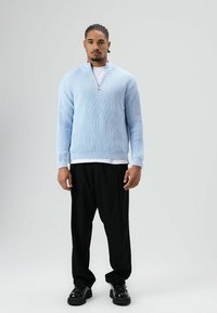 Pull-over bleu clair côtelé avec un col à demi-fermeture éclair, associé à un pantalon noir ample et des chaussures noires, créant un look décontracté.