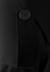 Vêtement en tissu noir avec une surface texturée, présentant un détail de bouton proéminent et un design en tissu plié le long du bord.