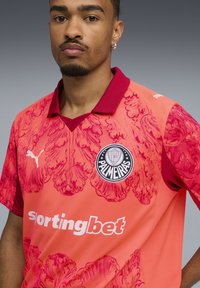 Camisa deportiva en rosa brillante con un patrón floral texturizado. Presenta un cuello marrón, un logo circular negro y el texto blanco "sportingbet."