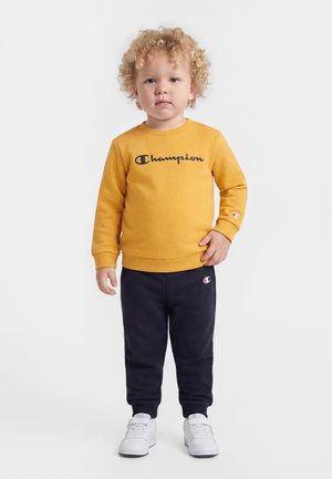 Ungt barn med krøllet hår iført en sennepsgul Champion-sweatshirt, marineblå joggingbukser og hvide sneakers, der står på en hvid baggrund.