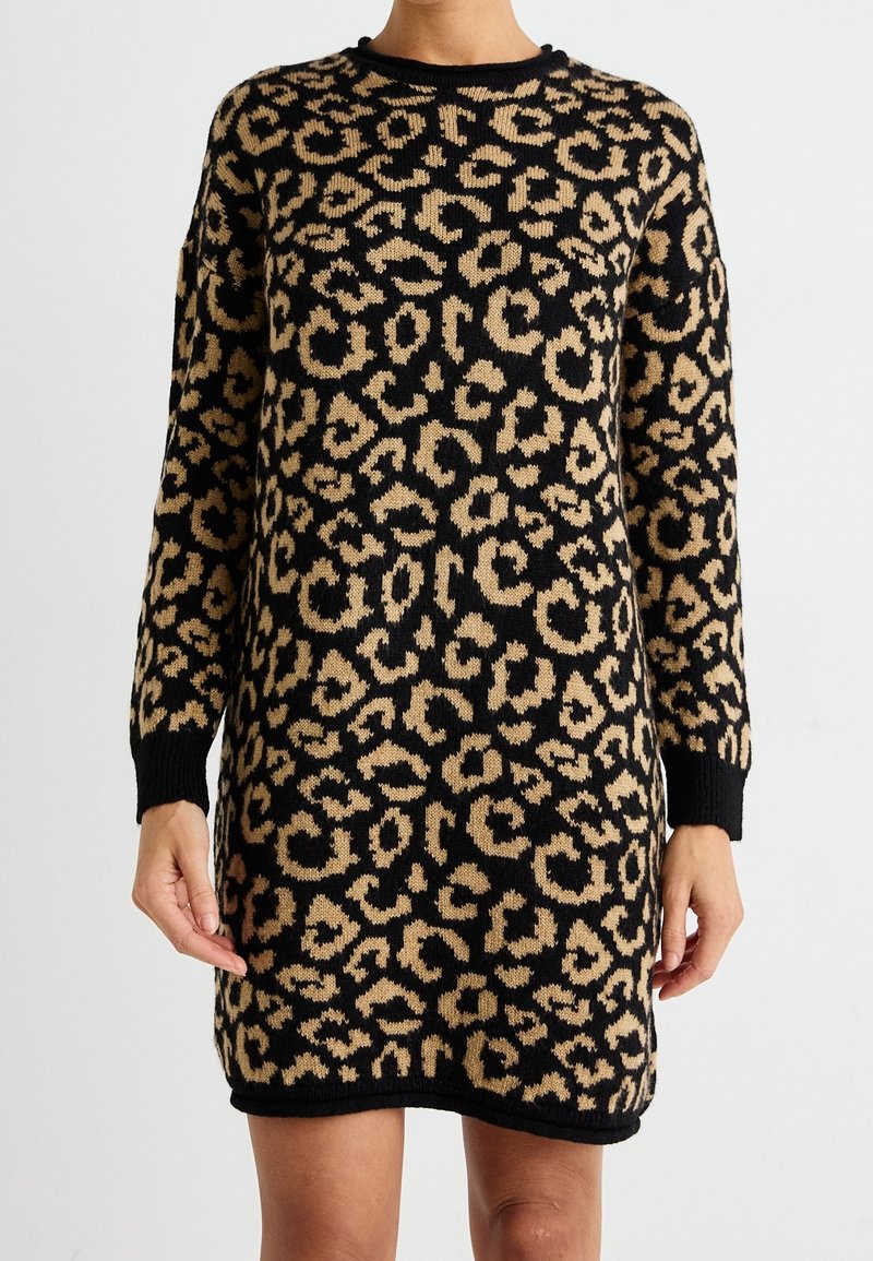 Femme portant une robe tricotée à manches longues, longueur aux genoux, avec un motif léopard noir et beige.