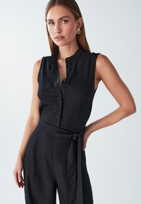 Črn brezrokavni kombinezon iz gladkega materiala, ki se ponaša z ovratnikom, sprednjimi gumbi in veznim pasom za poudarjen silhuet.