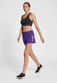 Schwarzer Sport-BH, lila Laufshorts mit pinken Chevron-Akzenten und schwarze Sneakers. Das Material wirkt leicht; ein elastischer Taillenbund ist sichtbar.