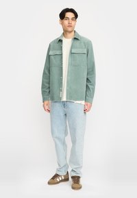 Lichtgroene ribfluwelen shirtjack met twee borstzakken, knoopsluiting, gelaagd over een licht top, gecombineerd met lichtblauwe jeans en bruine sneakers.