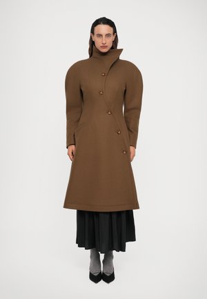 Een vrouw draagt een bruine asymmetrische knoopjas met hoge kraag, gecombineerd met een zwarte rok, grijze sokken en zwarte puntige schoenen op een witte achtergrond.
