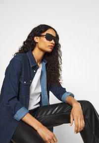 Occhiali da sole oversize neri, camicia di jeans in blu scuro e chiaro, t-shirt bianca, pantaloni in pelle neri, texture del tessuto lisce, vestibilità casual.