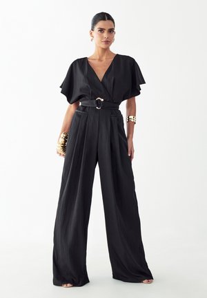 Kvinde står iført en sort jumpsuit med vide ben, V-hals, korte ærmer, bælte i taljen, gyldne armbånd og holder en gylden clutch.