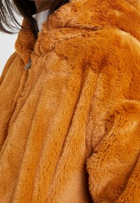 Oranžová mikina s umělou kožešinou a jemnou texturou, vybavená zipovým zapínáním a zaoblenou kapucí. Látka má lesklý, plush vzhled.